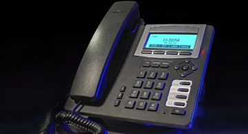 Schwarzes Bürotelefon mit Display, Tastenfeld, Funktionstasten und beleuchtetem Bildschirm auf dunklem Hintergrund.